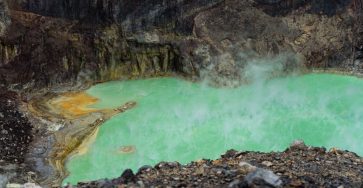 A steaming green acidic lake in a volcanic crater with vibrant color contrast against rocky terrain.