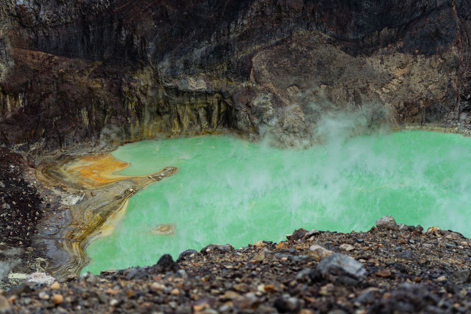 A steaming green acidic lake in a volcanic crater with vibrant color contrast against rocky terrain.